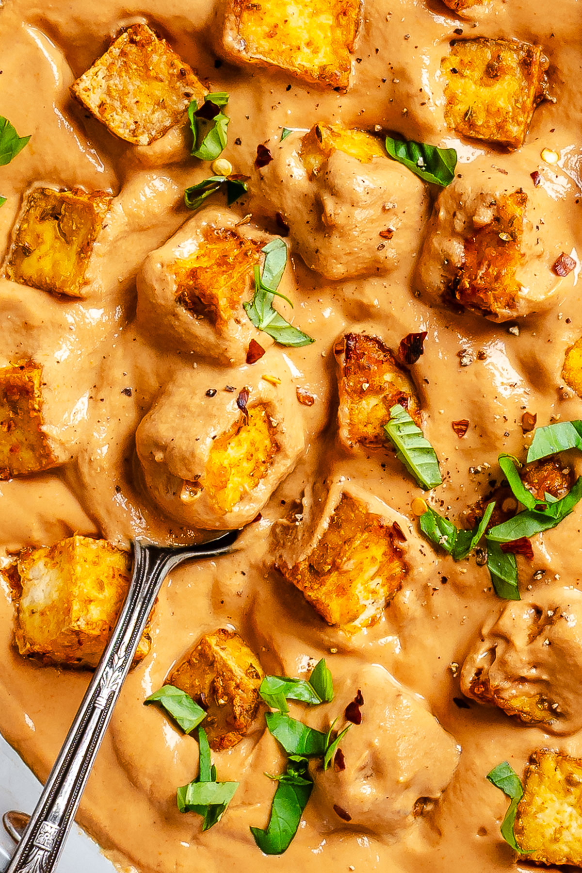 A spoon in a creamy brown sauce with golden tofu cubes, garnished with chopped green herbs and red pepper flakes.