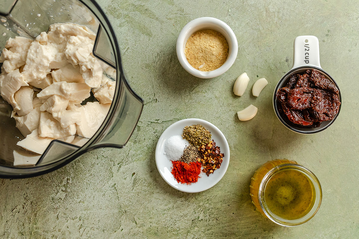 Ingredients for a sauce arranged on a countertop: tofu in a blender, chipotle peppers, olive oil, garlic cloves, nutritional yeast, and assorted spices on small plates.