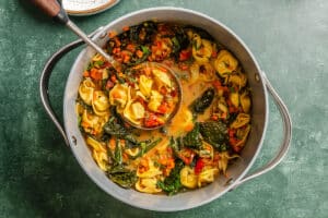 A pot of vegetable tortellini soup with leafy greens, diced carrots, and broth, with a ladle resting inside.