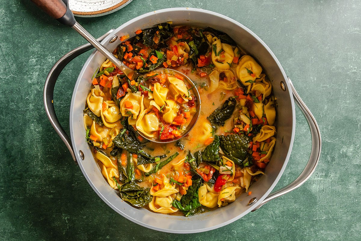 A pot of vegetable tortellini soup with leafy greens, diced carrots, and broth, with a ladle resting inside.