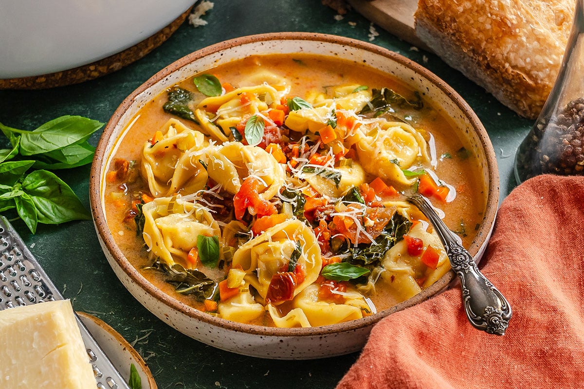 Bowl of tortellini soup with vegetables, topped with shredded cheese and basil, placed next to a grater, cheese wedge, and napkin.