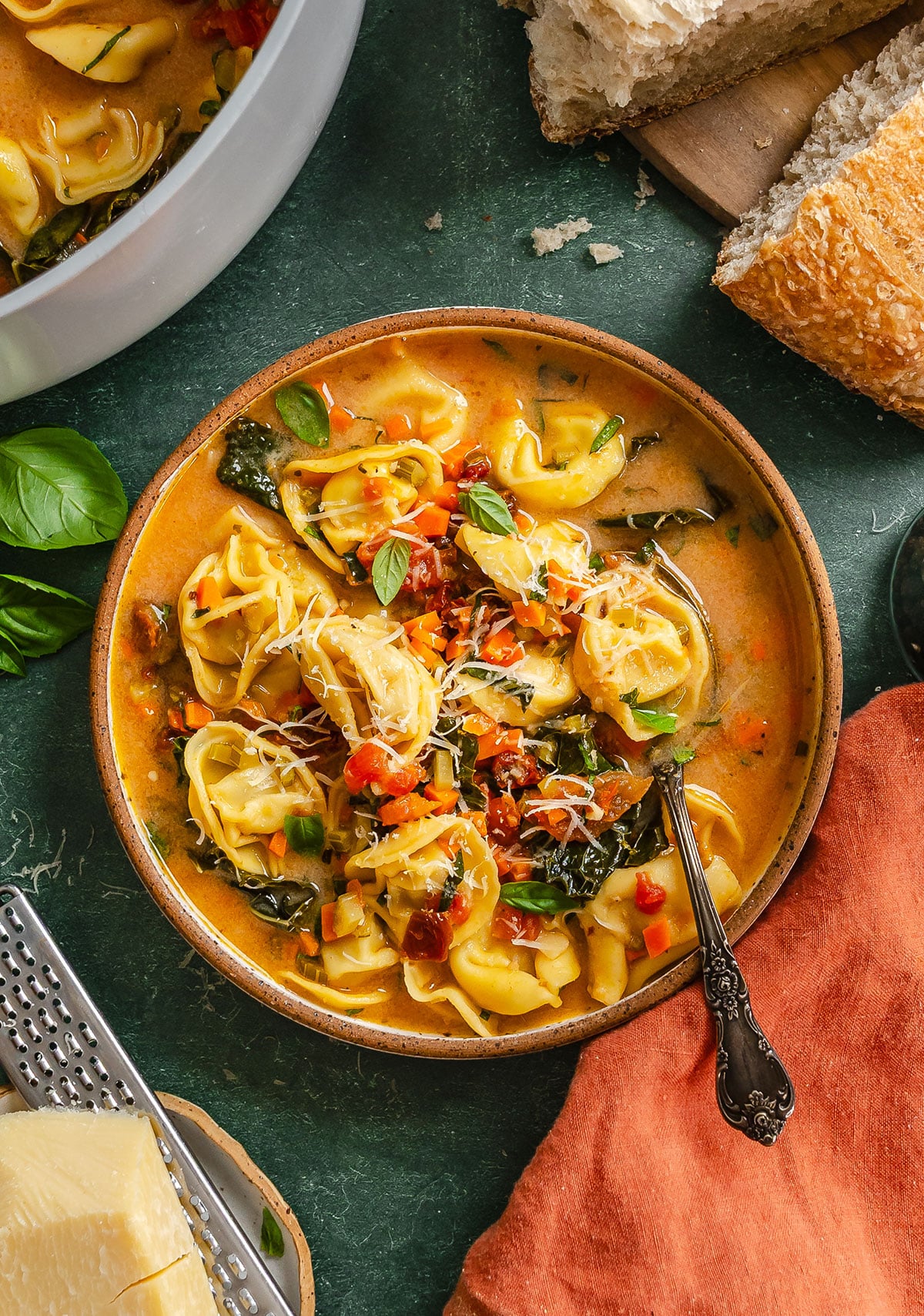 Bowl of tortellini soup with vegetables and greens, topped with shredded cheese and fresh basil, next to pieces of bread on a green surface.
