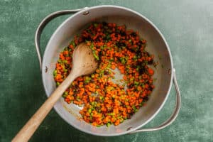 Diced carrots, celery, and onions being sautéed in a large pot with a wooden spoon on a green surface.