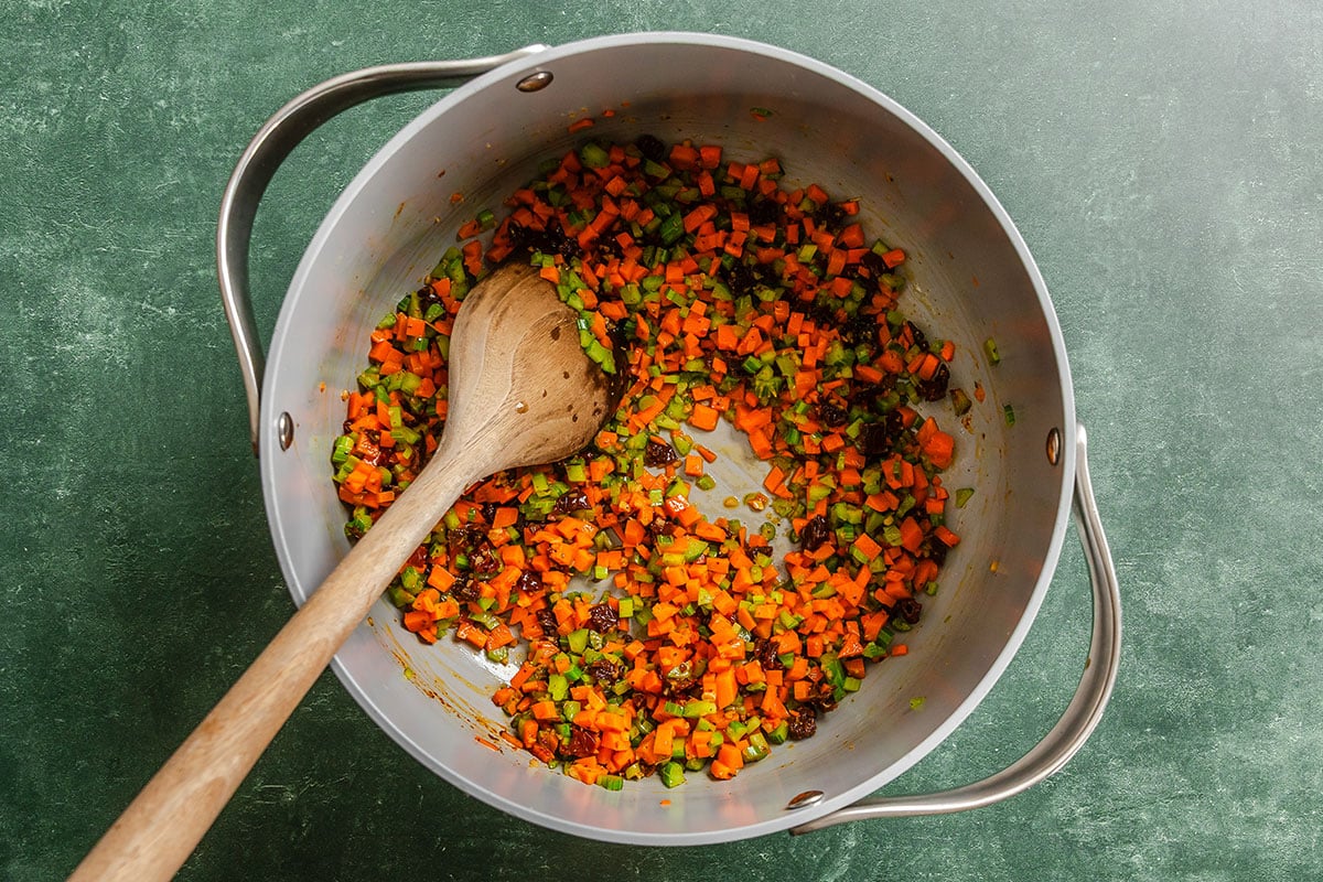 Diced carrots, celery, and onions being sautéed in a large pot with a wooden spoon on a green surface.