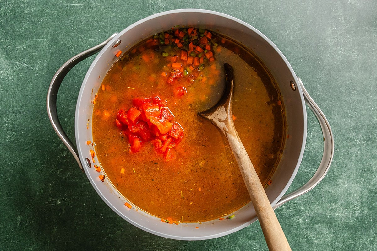A large pot with vegetable soup being stirred by a wooden spoon, topped with chopped tomatoes, sits on a green countertop.