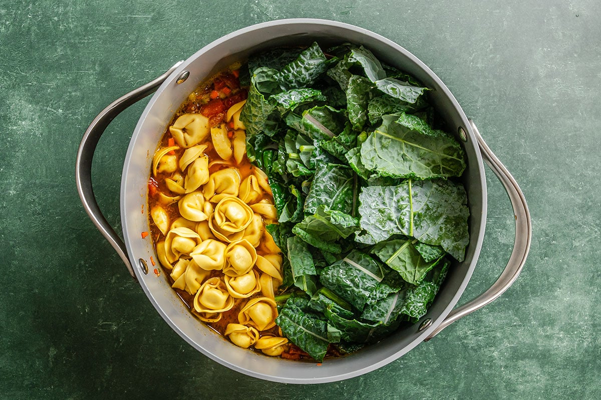 A pot filled with uncooked tortellini, chopped kale, and a tomato-based sauce, viewed from above on a green surface.