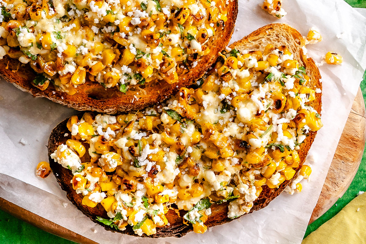 Two slices of toasted bread topped with grilled corn, crumbled cheese, herbs, and seasonings, served on parchment paper.