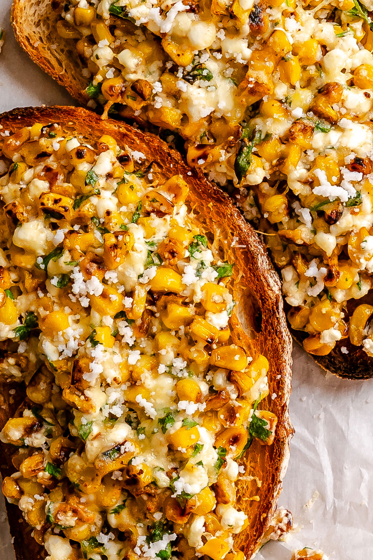 Toasted bread slices topped with roasted corn, melted cheese, herbs, and crumbled cheese on parchment paper.