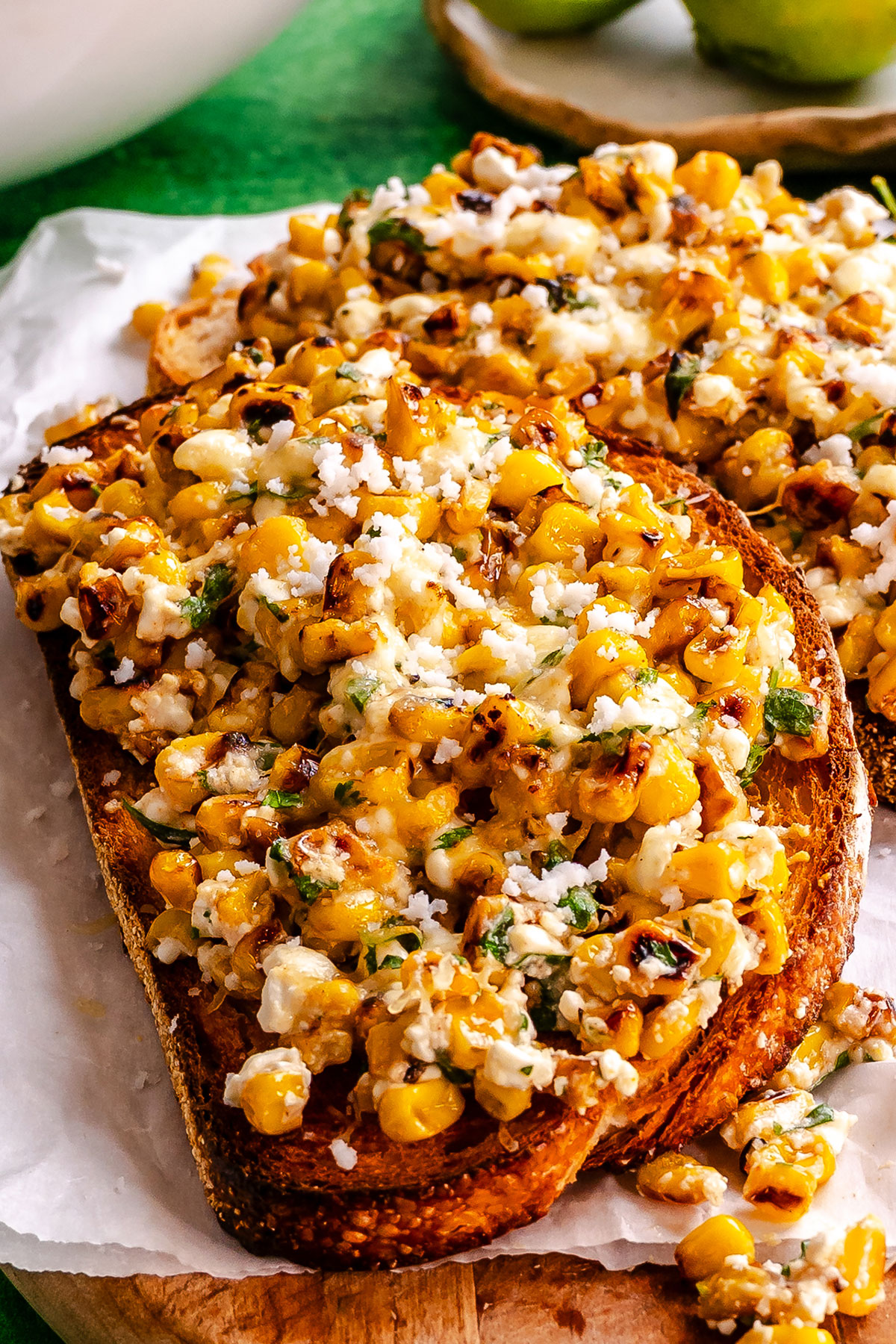 Two slices of toasted bread topped with roasted corn, herbs, and crumbled cheese are served on white parchment paper.