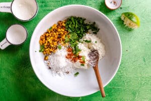 A white bowl contains corn, chopped cilantro, sour cream, grated cheese, and spices with a wooden spoon, on a green surface. Measuring cups, lime, and a small dish are nearby.