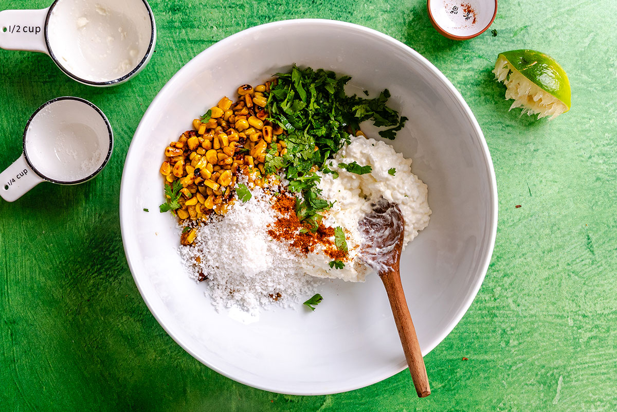 A white bowl contains corn, chopped cilantro, sour cream, grated cheese, and spices with a wooden spoon, on a green surface. Measuring cups, lime, and a small dish are nearby.