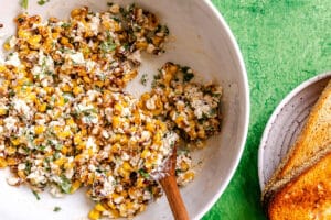 A bowl of creamy corn salad with herbs is mixed with a wooden spoon next to toasted bread on a white plate on a green surface.