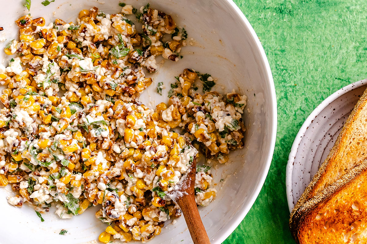 A bowl of creamy corn salad with herbs is mixed with a wooden spoon next to toasted bread on a white plate on a green surface.