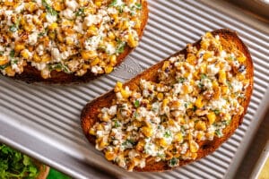 Two slices of toasted bread topped with a mixture of roasted corn, cheese, herbs, and spices sit on a ridged metal baking sheet.