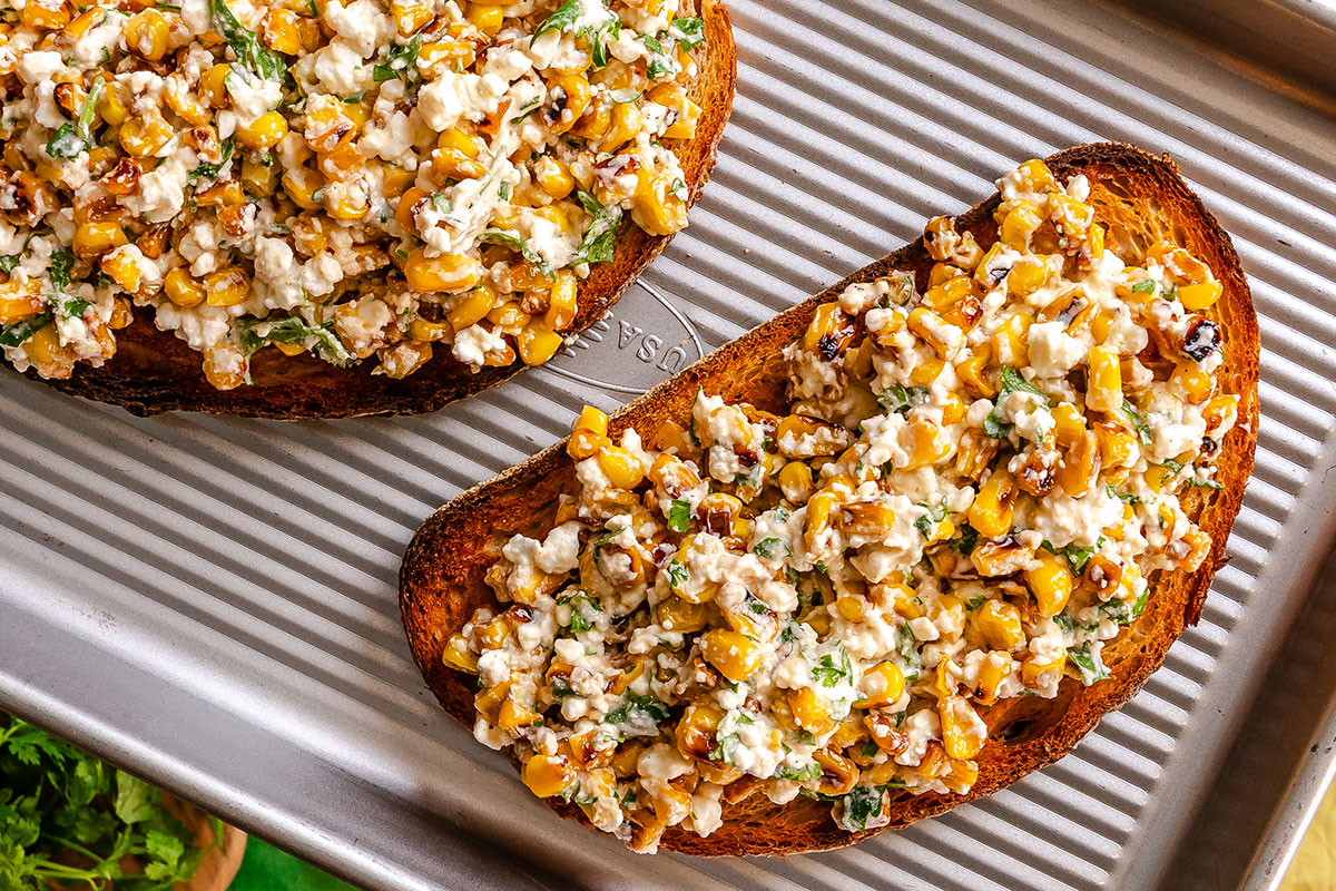 Two slices of toasted bread topped with a mixture of roasted corn, cheese, herbs, and spices sit on a ridged metal baking sheet.