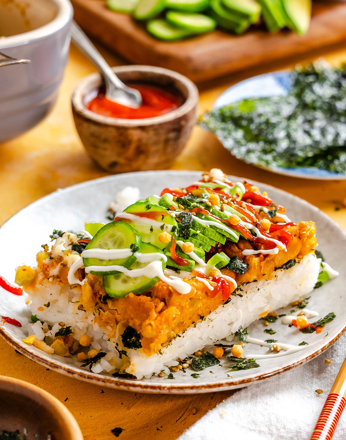 A plate with a sushi bake slice topped with sliced cucumber, avocado, sauces, and garnishes; nori sheets and sauce bowl are in the background.