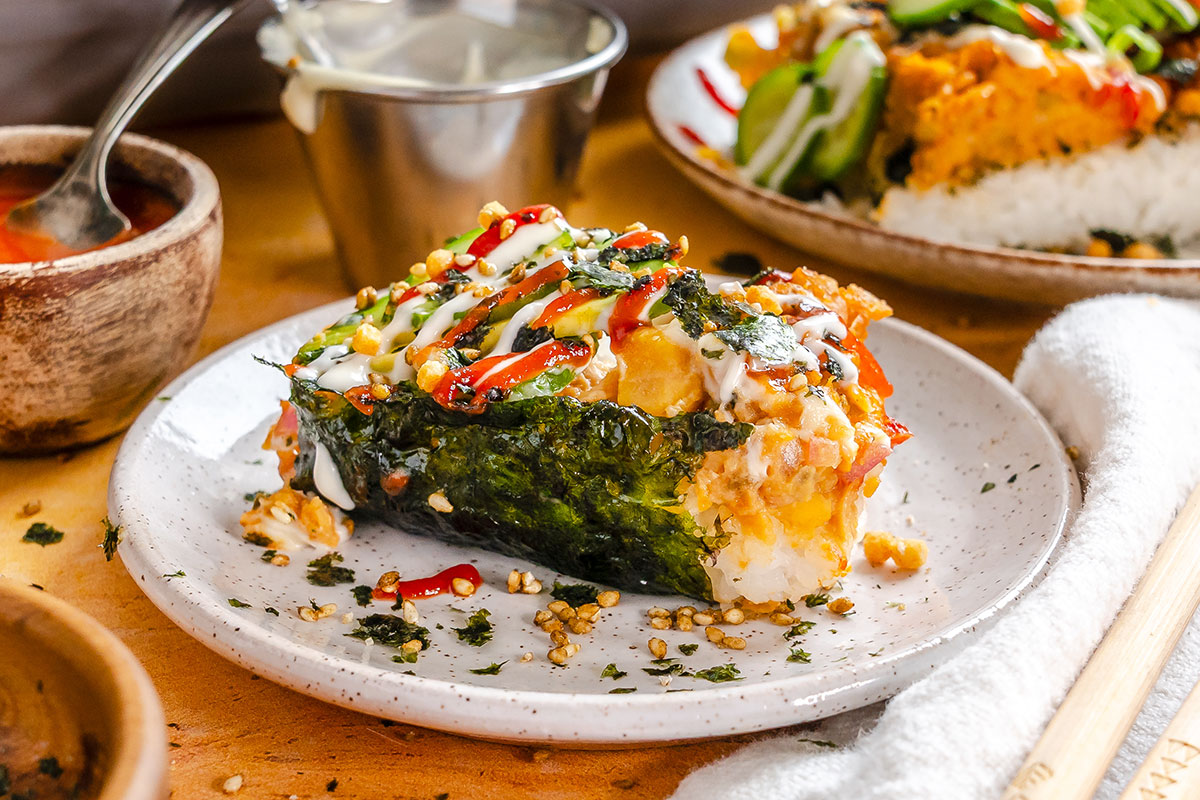 A slice of baked sushi casserole topped with sauces, nori, and sesame seeds sits on a speckled plate, surrounded by small bowls and chopsticks.