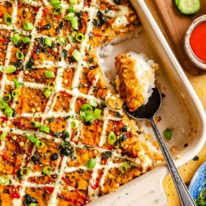 A baked casserole dish topped with chopped green onions, sauce, and mayonnaise, with a serving spoon and a square piece removed. Bowls of sauce, cucumber, and seaweed are nearby.