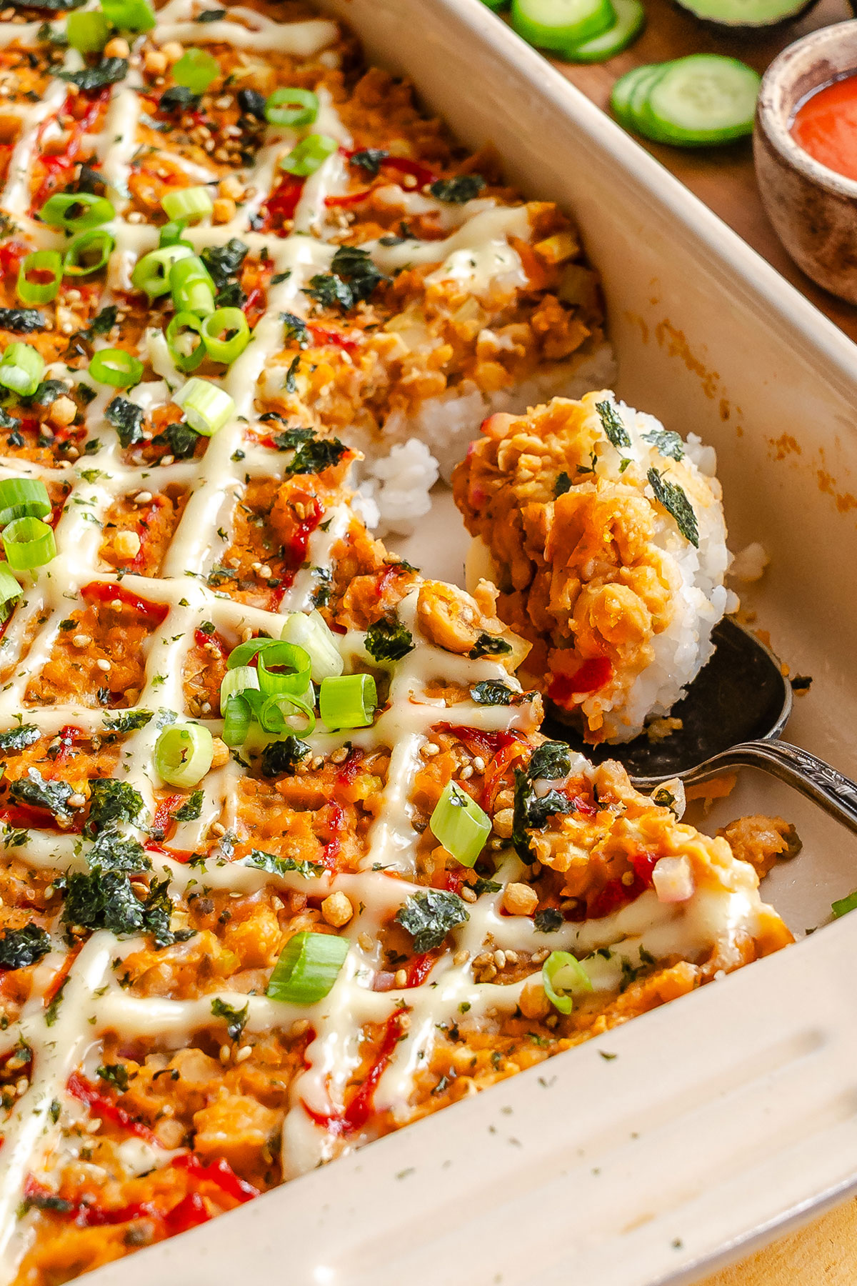 A casserole dish filled with a baked sushi casserole topped with sliced green onions, drizzled sauce, and herbs; a serving is being scooped out with a spoon.
