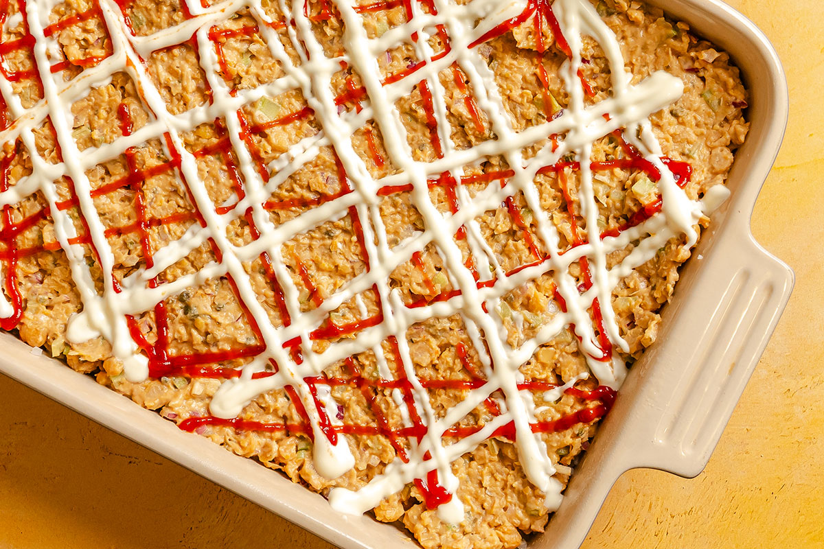 Baked casserole topped with white and red sauces drizzled in a crisscross pattern inside a rectangular baking dish on a yellow surface.