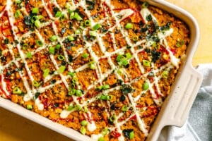 A baked casserole topped with zigzag lines of white and red sauces, chopped green onions, and crumbled seaweed in a rectangular dish.