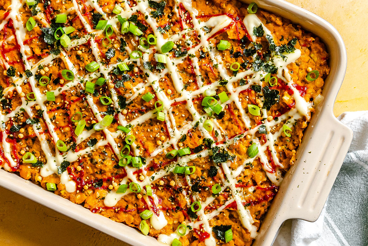 A baked casserole topped with zigzag lines of white and red sauces, chopped green onions, and crumbled seaweed in a rectangular dish.