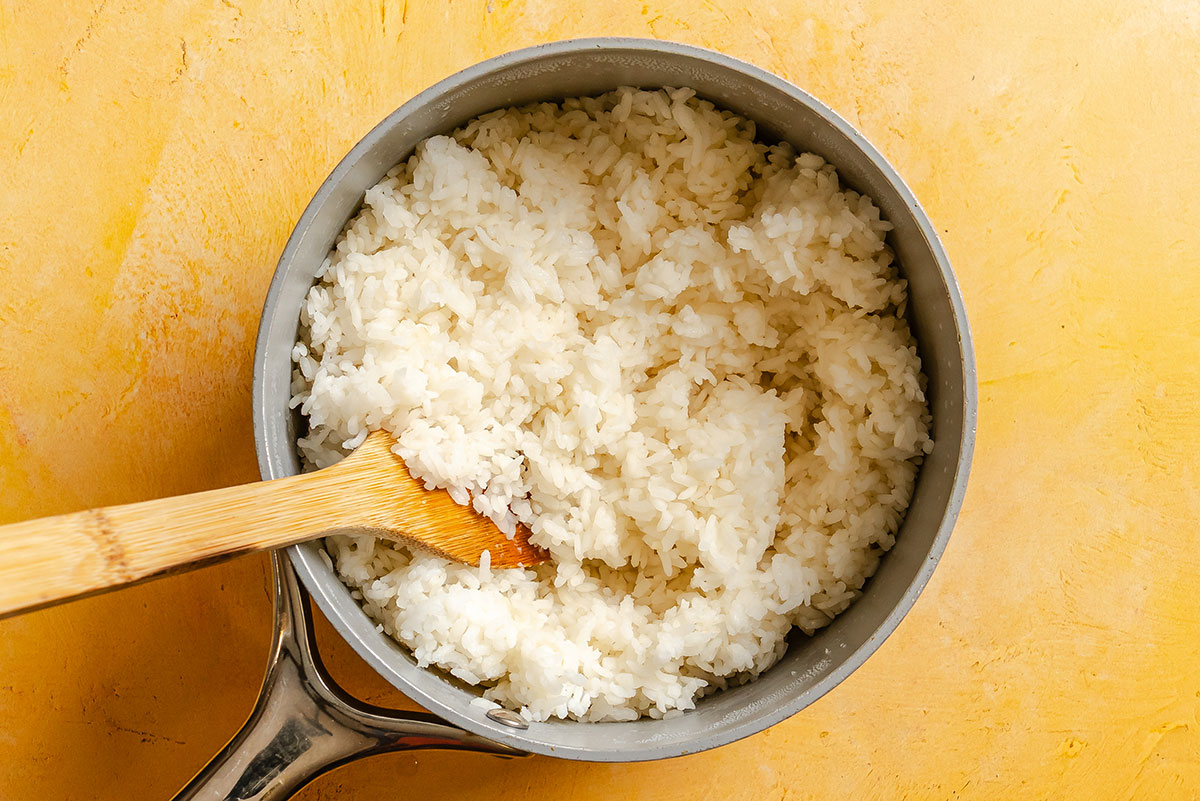 A pot of cooked white rice with a wooden spoon resting inside, placed on a yellow surface.