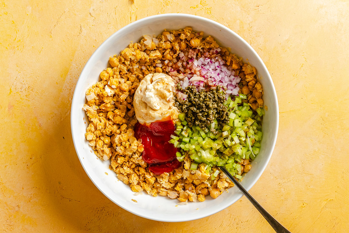 Bowl containing mashed chickpeas, chopped celery, red onion, capers, mayonnaise, and ketchup, with a spoon on the side, on a yellow background.