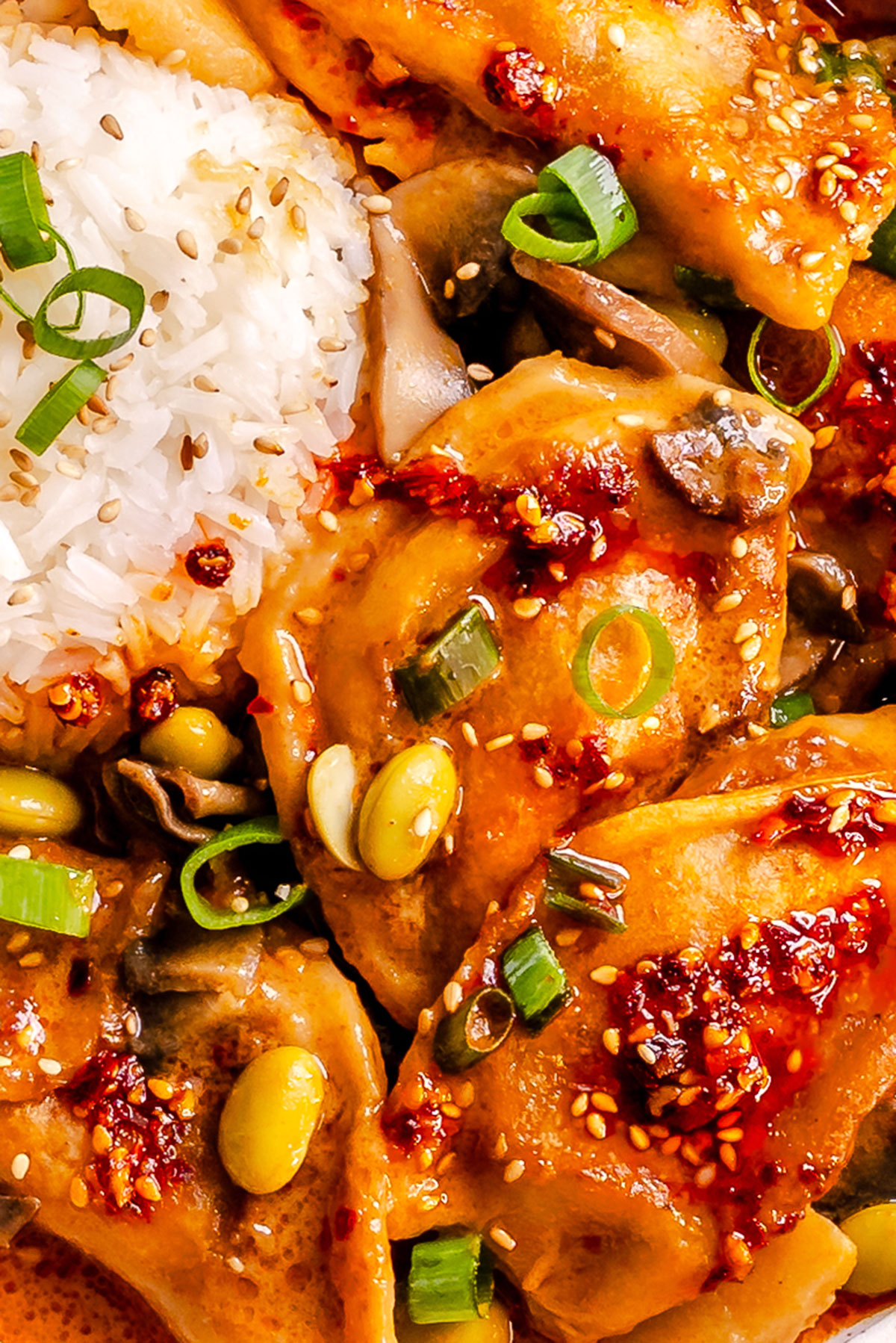 Close-up of dumplings topped with chili sauce, green onions, and sesame seeds, served with white rice, edamame, and mushrooms.