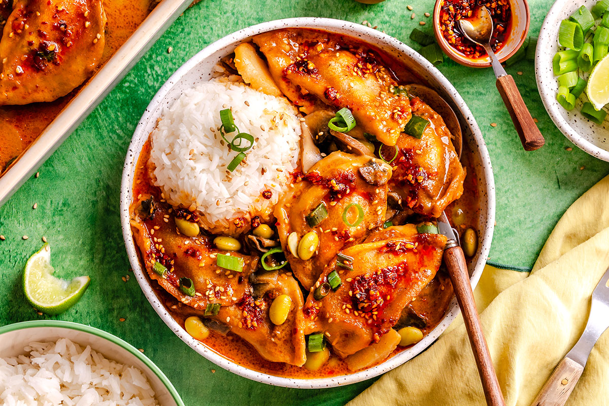 Bowl of rice and saucy dumplings garnished with green onions and sesame seeds, served with a spoon. Lime wedge and extra rice are on the side.
