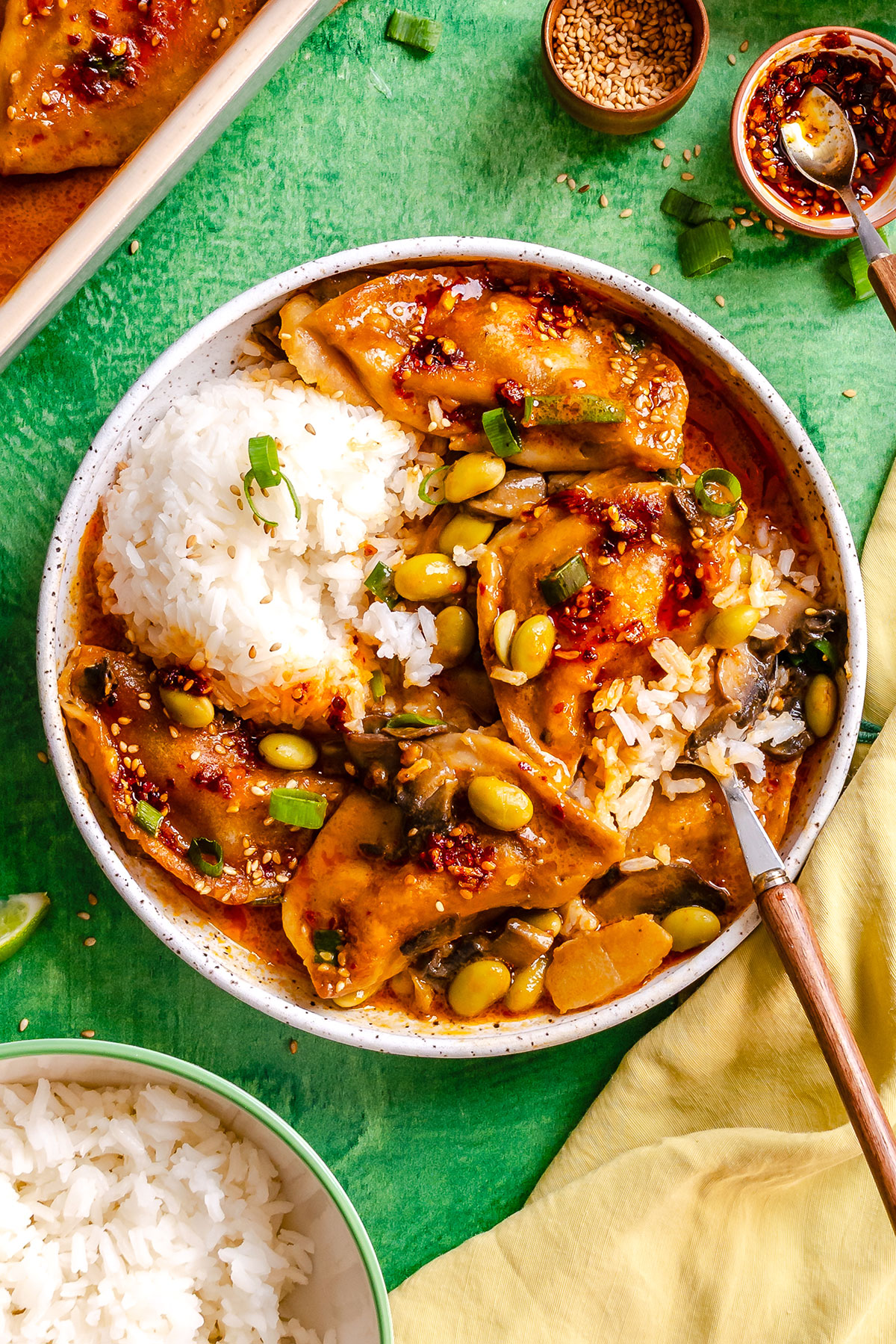 A bowl of white rice with saucy dumplings, edamame, vegetables, and chili oil, garnished with green onions, on a green surface with extra rice, sesame seeds, and sauce nearby.