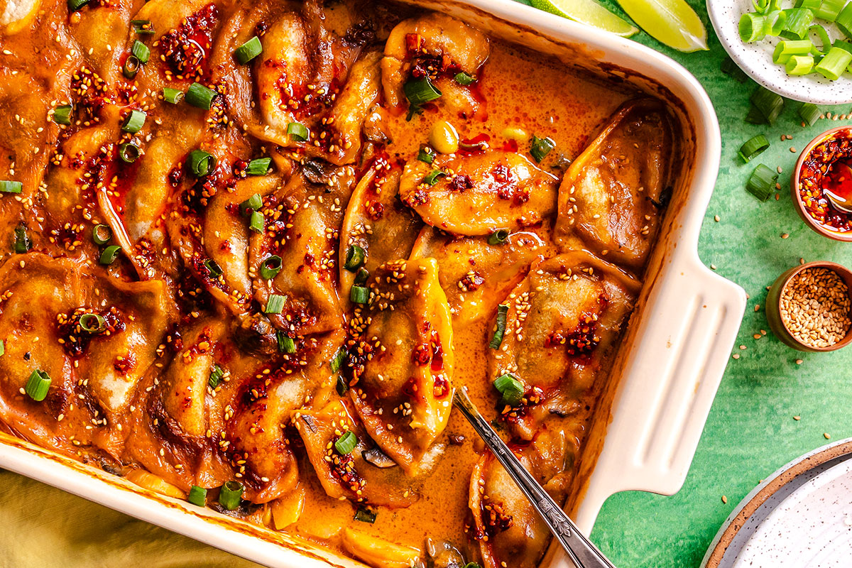 A baked dish of dumplings in a spicy, saucy glaze, topped with chopped green onions and sesame seeds, with a spoon in the corner. Lime wedges and condiments are visible on the side.
