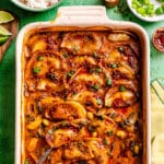 Baked dumplings in a rectangular dish, topped with a spicy red sauce, sliced green onions, and sesame seeds, with a spoon on the side and small bowls of rice and garnishes nearby.