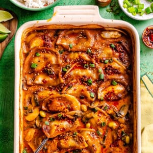 Baked dumplings in a rectangular dish, topped with a spicy red sauce, sliced green onions, and sesame seeds, with a spoon on the side and small bowls of rice and garnishes nearby.