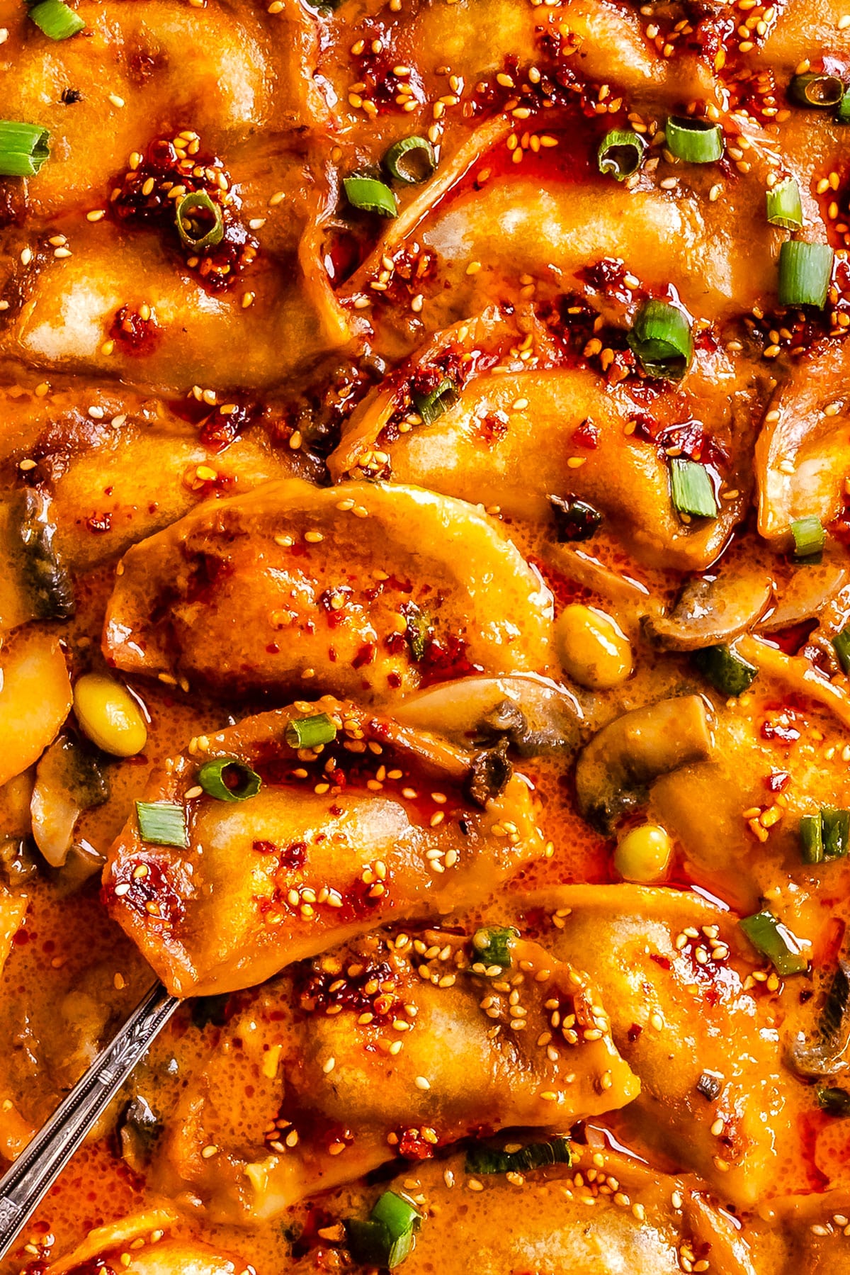 Close-up of cooked dumplings in a spicy red sauce, garnished with chopped green onions and sesame seeds.