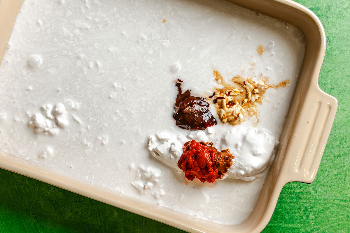 A beige baking dish contains coconut milk with separate dollops of red curry paste, brown sauce, minced garlic, and grated ginger on top, set on a green surface.