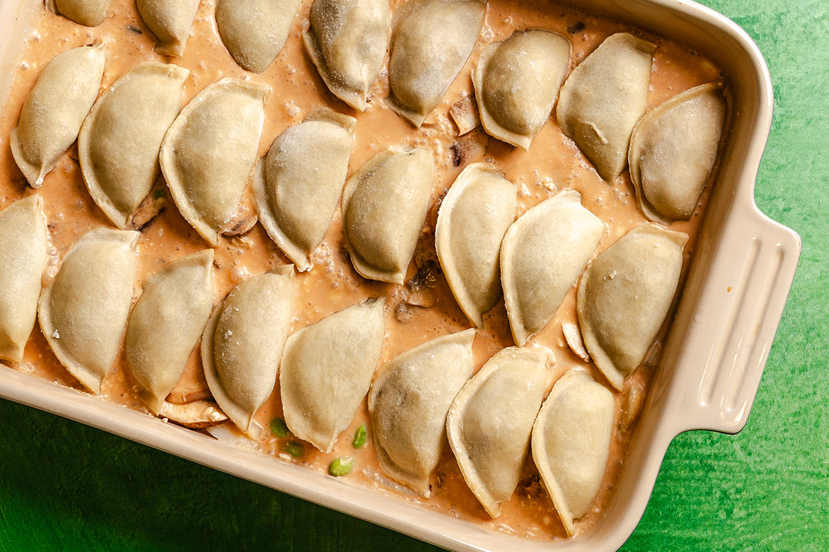 Rectangular casserole dish filled with pierogies arranged in rows over a creamy sauce, shown on a green surface.