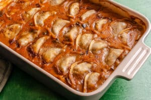 Rectangular baking dish filled with cooked dumplings or pierogi in a thick, reddish-brown sauce, arranged in overlapping rows on a green surface.