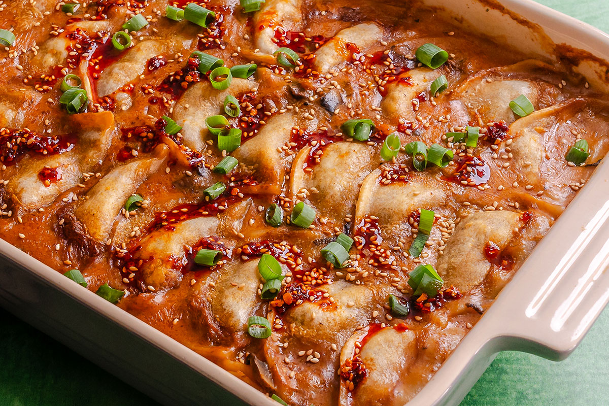 A baked casserole dish filled with dumplings in a reddish sauce, garnished with sliced green onions, sesame seeds, and chili oil.