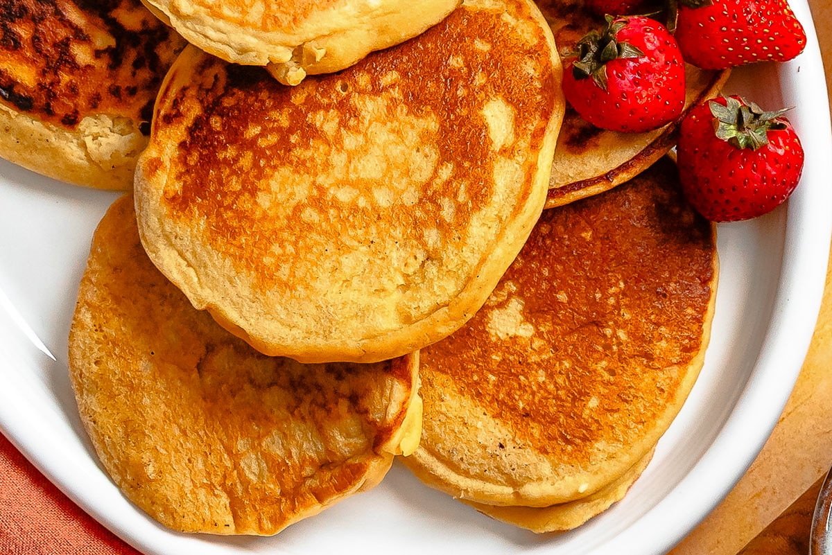 A close-up of golden brown pancakes stacked on a white plate, with fresh strawberries placed on the side.