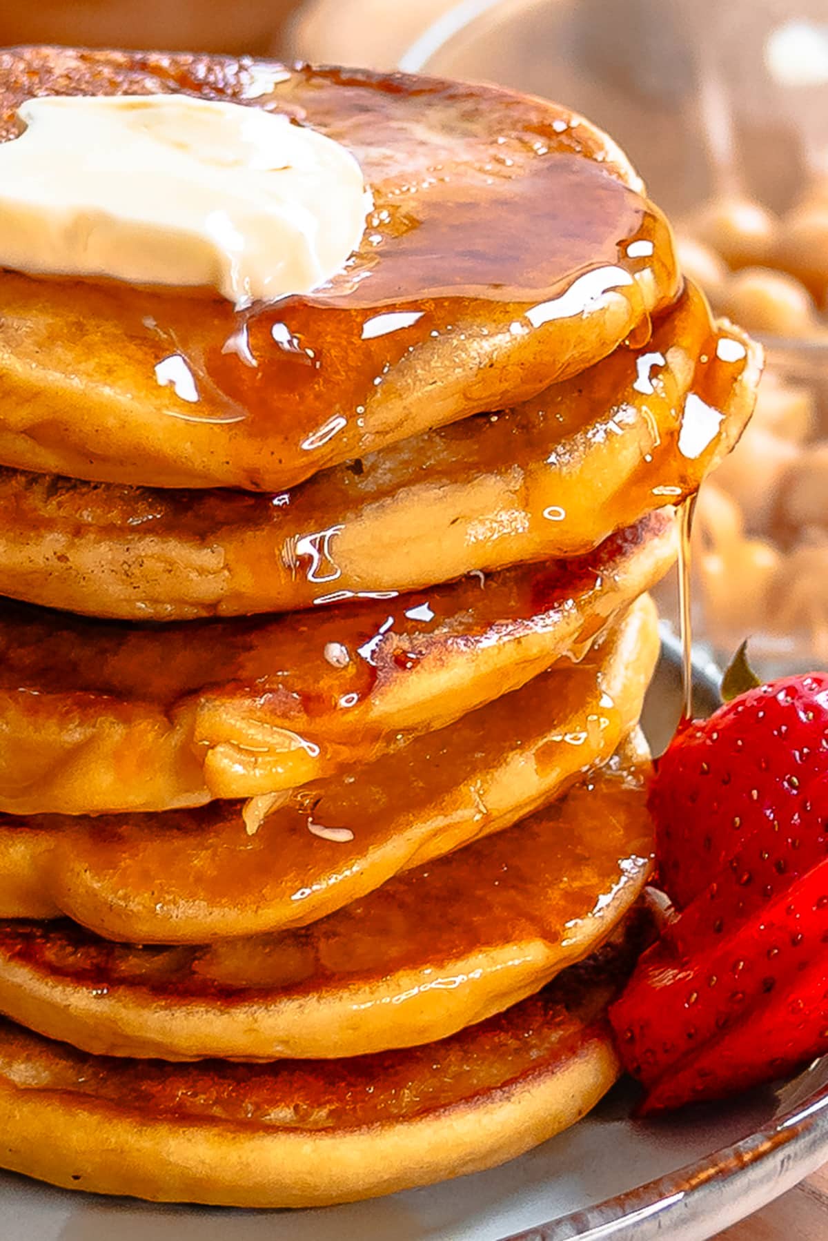A stack of pancakes topped with butter and syrup sits next to sliced strawberries and a bowl of nuts.