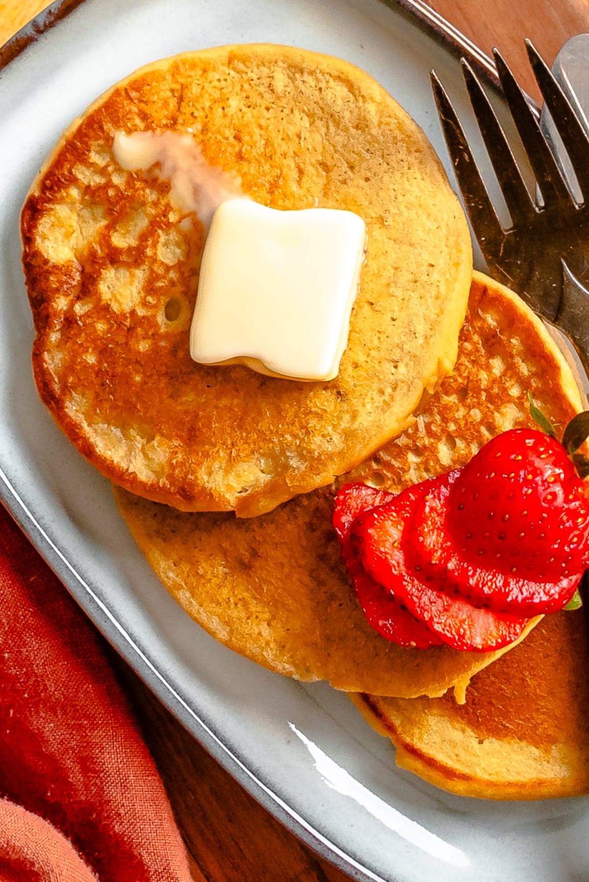 Two golden chickpea pancakes topped with a pat of butter and sliced strawberries are served on a white plate with a fork nearby.
