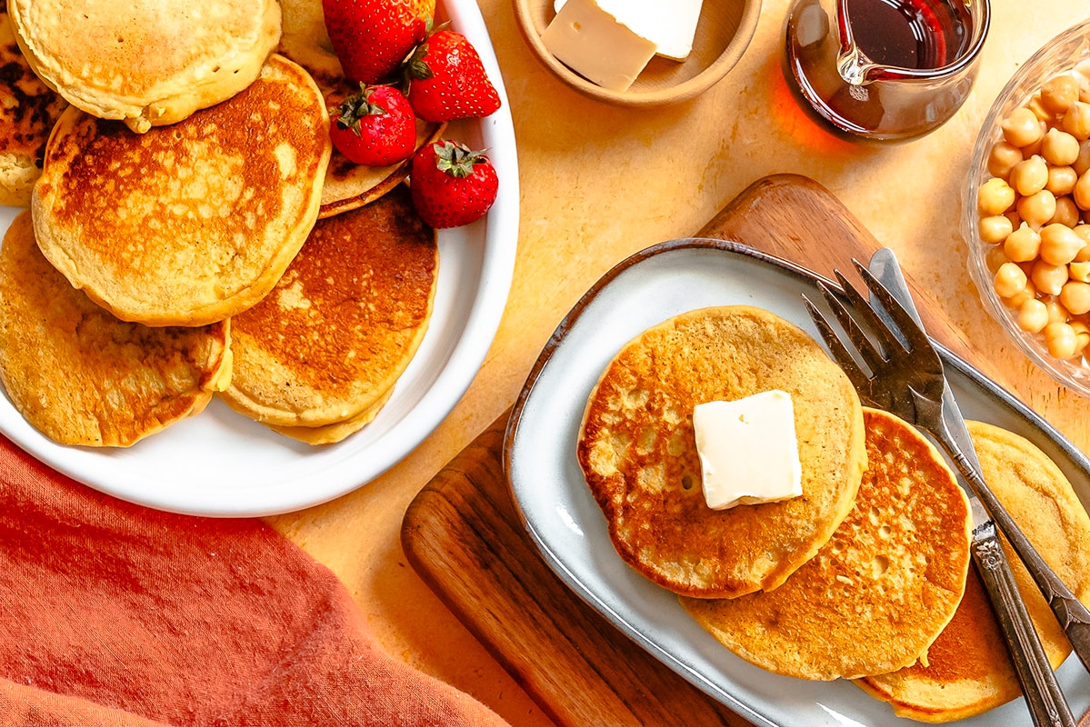 Plate of pancakes with a pat of butter, strawberries, syrup, and a bowl of chickpeas on a table with an orange napkin.