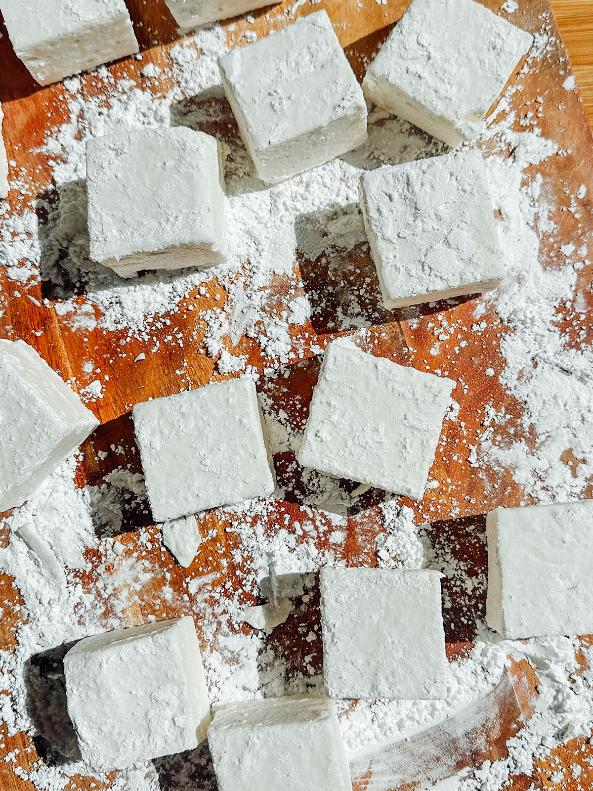 Cubes of homemade marshmallows coated in powdered sugar are arranged on a wooden surface dusted with more sugar.