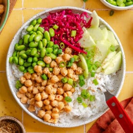 A bowl with white rice, chickpeas, edamame, sliced cucumber, shredded red cabbage, green onions, and a fork, on a yellow tiled surface.