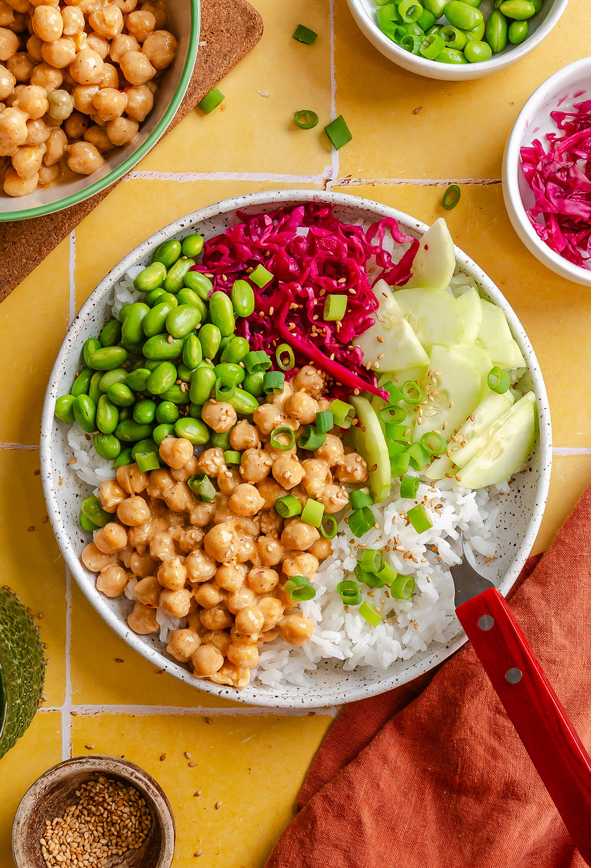 A bowl with white rice, chickpeas in sauce, edamame, sliced cucumber, and shredded red cabbage, garnished with green onions. A fork is on the plate; small bowls of toppings are nearby.
