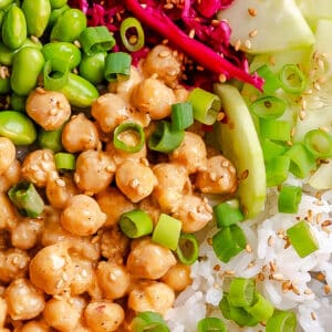 A close-up of a colorful bowl with white rice, chickpeas in sauce, edamame, shredded red cabbage, cucumber slices, green onions, and sesame seeds.