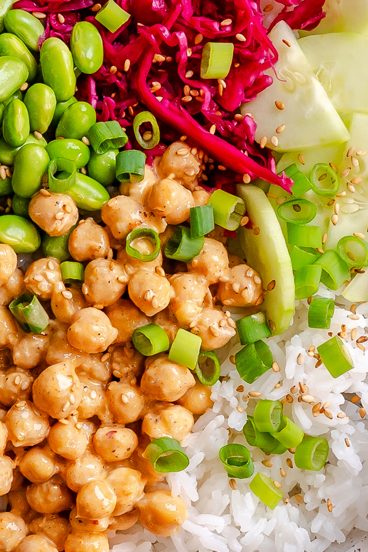A close-up of a colorful bowl with white rice, chickpeas in sauce, edamame, shredded red cabbage, cucumber slices, green onions, and sesame seeds.