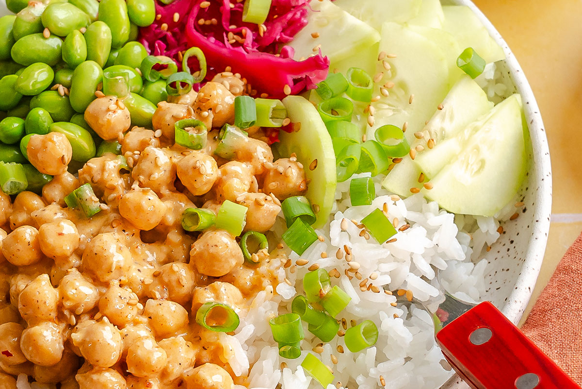 A bowl with rice, sliced cucumber, chickpeas in sauce, edamame, pickled cabbage, green onions, and sesame seeds, with a red-handled fork.