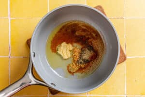 A gray pan on a yellow tiled surface contains oil, minced garlic, ginger paste, and various ground spices.